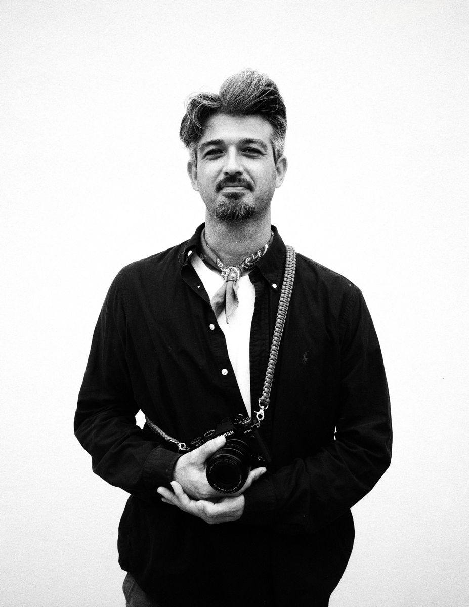 Hajime Birnbaum in a black shirt and bandana, holding his Fujifilm camera, in black and white.