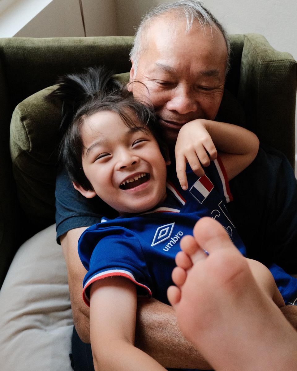 A boy in a blue soccer jersey laughs with his eyes squeezed shut while his grandfather hugs him from behind in a green armchair.
