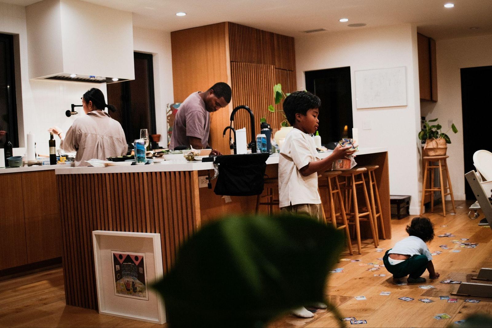 A wide view of an open kitchen. Parents cook at the counter while one child stands at the island and the youngest sits on the wood floor.